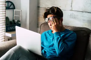 A young man wearing glasses and a blue long-sleeve shirt sits on a couch with his laptop, looking tired and holding his head.