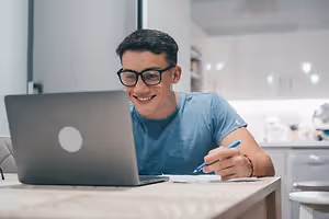 A man is smiling and seems to be working on his laptop while sitting on a chair in front of a table in the kitchen.