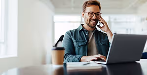 A smiling man wearing glasses is talking on his cellphone while working on his laptop at his desk in an office room.