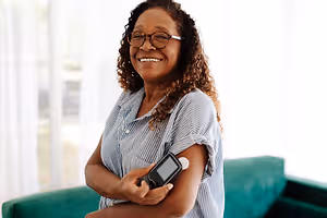 An adult woman with curly hair wearing glasses, smiling while holding a blood glucose meter on her arm.