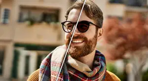 A man with a beard wearing sunglasses and a plaid scarf is smiling and standing outside a building.