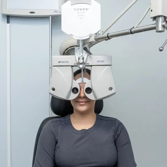 A woman is sitting in front of an eye exam machine with her eyes closed