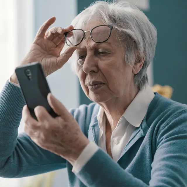 Senior woman with white hair holding a smartphone and wearing glasses, looking at the camera, and adjusting her glasses