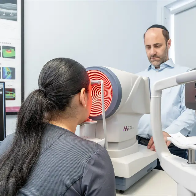 A woman is undergoing an eye examination with a red light in the background, while a man stands behind her, observing. Both are in a room with a large monitor displaying images and text on the wall behind them.