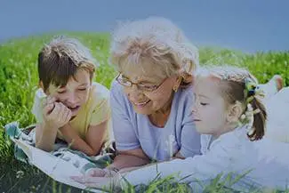 An older woman with her grandchildren lying on a blanket and reading a book together in a grassy field