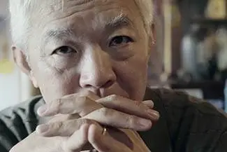 An older man with white hair wearing a black shirt is holding a stick in his mouth while looking at something
