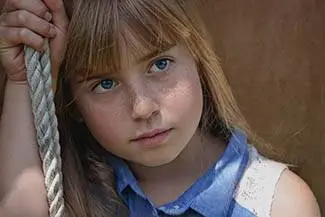 A young girl with blonde hair and blue eyes holds a rope with her left hand while looking at the camera.