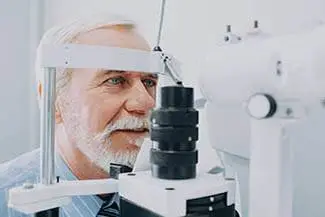 An older man with a white beard and glasses looking through an eye exam machine