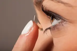 A woman wearing white nail polish is inserting a contact lens into her left eye with her left hand.