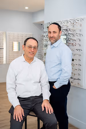 Two men in an eye care store. One is wearing glasses and the other is wearing a yarmulke. They are standing in front of a wall full of eyeglasses.