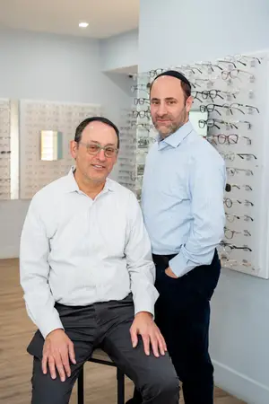 Two men in an eye care store. One is wearing glasses and the other is wearing a yarmulke. They are standing in front of a wall full of eyeglasses.