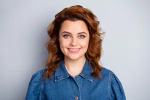 A smiling young woman with curly hair and blue eyes wearing a denim shirt against a white background.