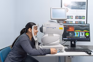 A woman is sitting at a desk, smiling and looking into an eye examination machine while wearing a gray shirt.