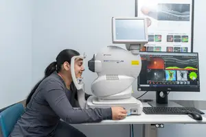 A woman is sitting at a desk, smiling and looking into an eye examination machine while wearing a gray shirt.