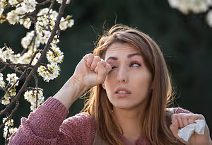 A woman touching her eye while standing under a tree with white flowers in the background
