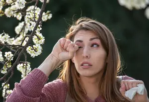 A woman touching her eye while standing under a tree with white flowers in the background