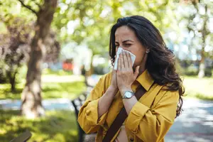 A woman wearing a yellow shirt and a watch is blowing her nose with a tissue in a park.