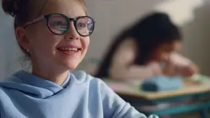 A smiling girl wearing glasses sits at a desk in a classroom with another student in the background.