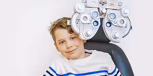A young boy with a smiling face sitting on a chair behind a phoropter