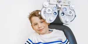 A young boy with a smiling face sitting on a chair behind a phoropter