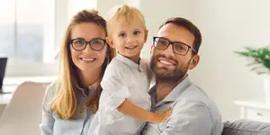 A family of three is smiling.The man and woman is wearing glasses, while the child are not. They are sitting on a couch