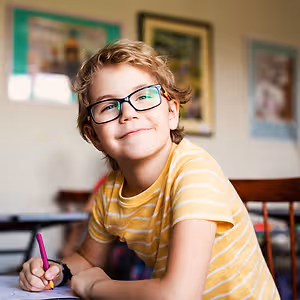 A smiling boy wearing glasses is sitting on a chair and holding a pen in his hand while writing on a piece of paper