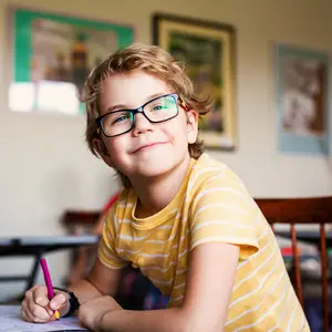 A smiling boy wearing glasses is sitting on a chair and holding a pen in his hand while writing on a piece of paper