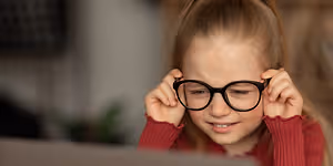 A young girl is wearing black glasses and a red sweater while she looks at the laptop