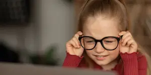 A young girl is wearing black glasses and a red sweater while she looks at the laptop