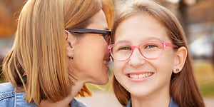 A young girl wearing pink glasses stands beside a woman with short blonde hair, both smiling and looking at each other in a park-like setting with cars and trees in the background
