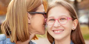 A young girl wearing pink glasses stands beside a woman with short blonde hair, both smiling and looking at each other in a park-like setting with cars and trees in the background