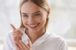 A woman wearing a white shirt is holding a white container of contact lens solution in her hands.