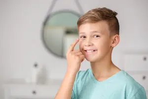 A young boy is smiling and pointing to his eye with his index finger.