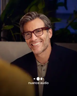 A smiling man in a suit and glasses looking forward, with a plant in the background and a watermark that reads Nuance Audio