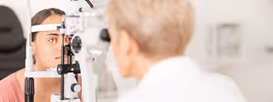 A young woman with brown eyes having her eyes examined by an optometrist in a white coat using a slit lamp.