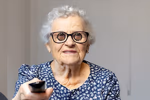 Elderly woman wearing glasses and holding a remote control while sitting on a chair.