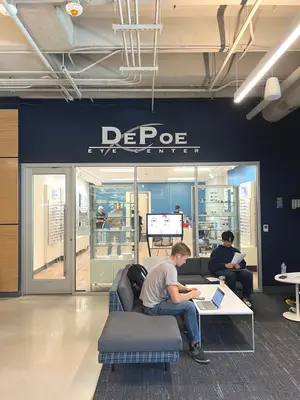 Two people are sitting on a couch inside the DePoe Eye Center with a laptop, a monitor, and a bottle of water on a table in front of them. The shop has glass doors with the logo of DePoe Eye Center. A man is standing inside the shop, and another man is sitting on the couch. There are two people standing in the distance, and there are several lights hanging from the ceiling.