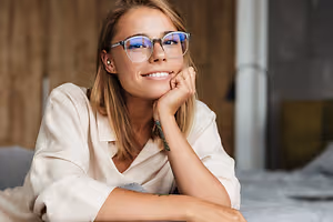 A Stockbridge woman in glasses smiles while sitting on a bed with her hand on her chin.