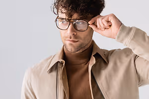 A man wearing glasses and adjusting them with one hand while wearing a turtleneck shirt and a beige jacket.