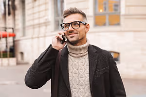 A Stockbridge  man in a black coat and glasses is talking on a cellphone on a city street
