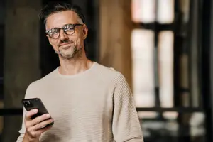 A smiling man holding a cell phone and wearing glasses in a room with glass windows.