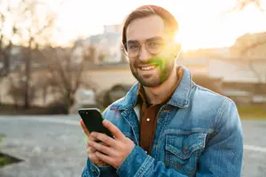 Man standing outside with a cellphone smiling and wearing glasses
