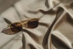 A pair of gold-framed sunglasses with brown lenses is placed on a beige silk cloth, which is folded in half and has a smooth texture in display oin Stockbridge.