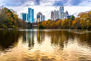 A city skyline with high-rise buildings, reflected on the water, surrounded by trees and a cloudy sky