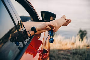 A person with their feet up on the car window and sunglasses in their hand.