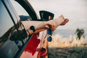A person with their feet up on the car window and sunglasses in their hand.