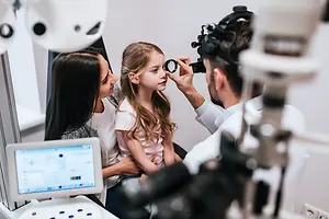 A young girl is having her eyes checked by an eye doctor with her mom standing by her side.