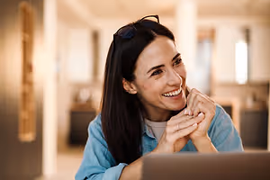 A woman is smiling and looking at a laptop in an indoor room with a white wall and a window.