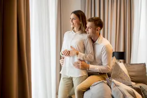 A couple sitting on a couch with pillows, smiling and looking out the window