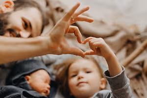 A man and his two children are lying on a bed and are making a heart shape with their hands. The child on the right is wearing a hat, while the child on the left is sleeping. The man is looking at them.
